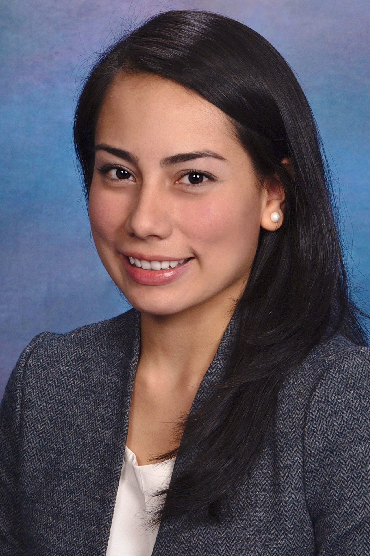 Portrait of a smiling young woman with long dark hair, wearing a gray blazer and white blouse, against a blue studio backdrop.